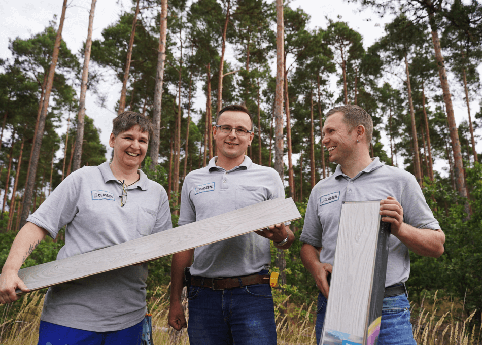 Trzech CLASSEN w szarych koszulkach polo z logo firmy stoi przed lasem, uśmiechając się i trzymając w rękach ekologiczne wykładziny podłogowe. Zdjęcie symbolizuje produkcję przyjazną dla środowiska, pracę zespołową oraz połączenie natury i nowoczesnej produkcji.