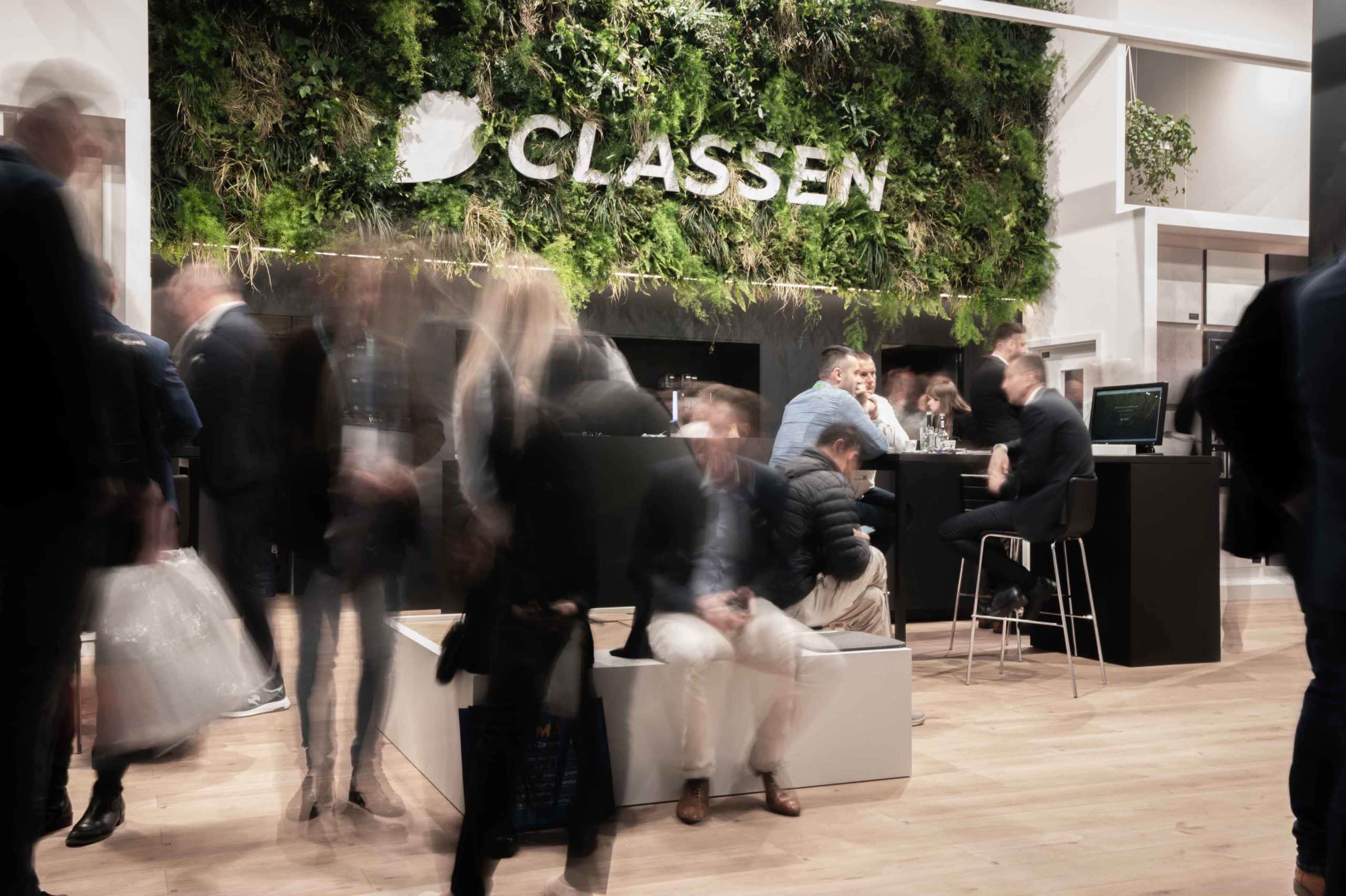 Goście odwiedzający CLASSEN z efektowną zieloną ścianą roślinną i nowoczesnym designem. Ludzie rozmawiający i wymieniający się opiniami na temat zrównoważonych wykładzin podłogowych i innowacji firmy CLASSEN.