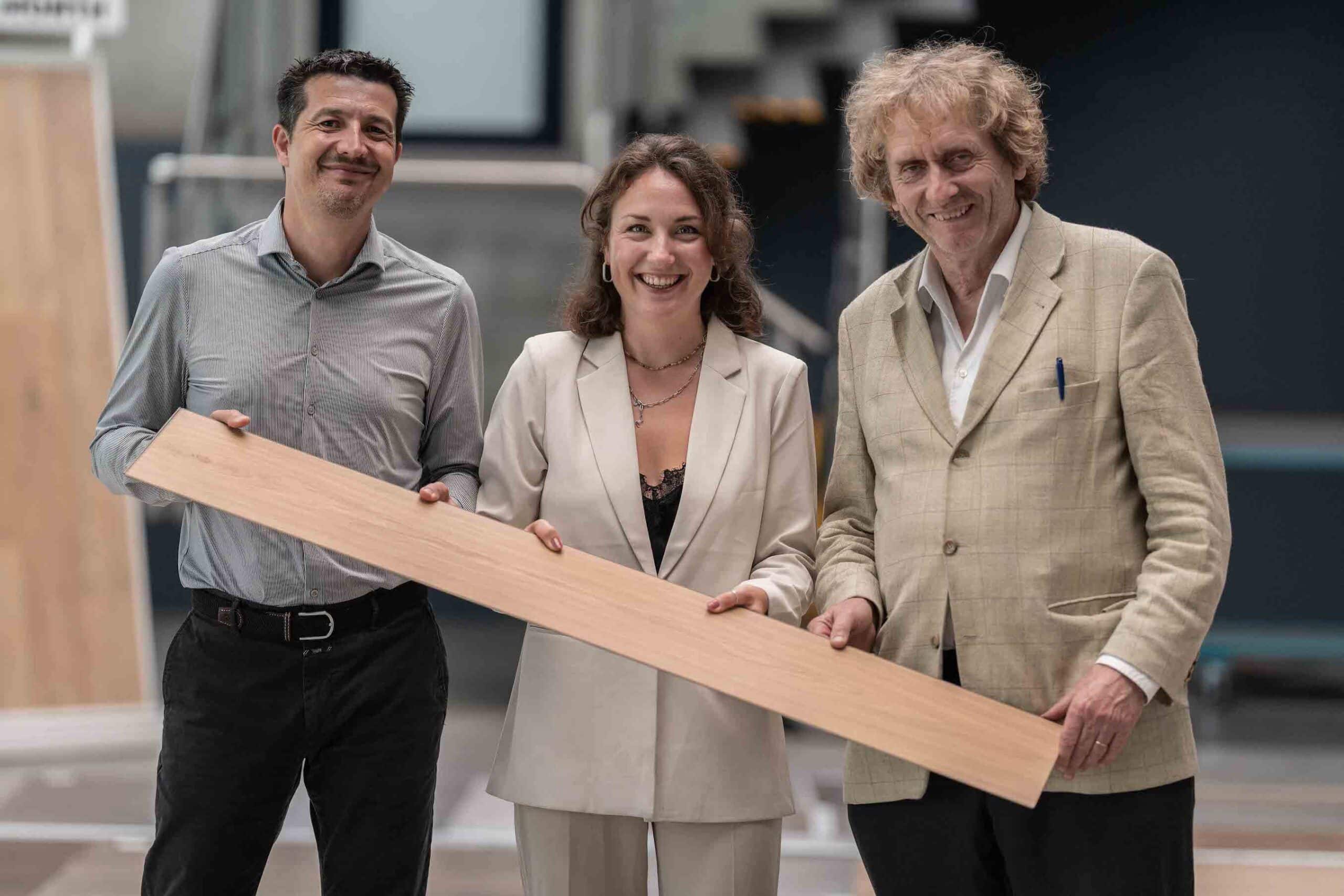 Trzy osoby trzymają wspólnie długi panel podłogowy i uśmiechają się do aparatu – symbol pracy zespołowej, innowacyjności i zrównoważonego rozwoju podłóg w CLASSEN.