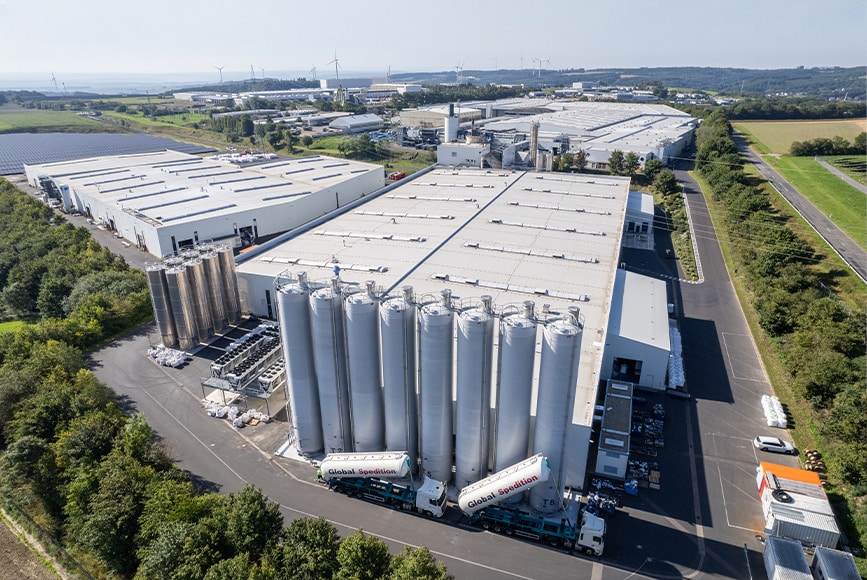 Zdjęcie lotnicze CLASSEN z dużymi silosami przemysłowymi, halami produkcyjnymi i przyległą infrastrukturą, otoczonymi zielonym krajobrazem i wiatrakami – symbolem nowoczesnej, zrównoważonej i oszczędzającej zasoby produkcji podłóg w Niemczech.