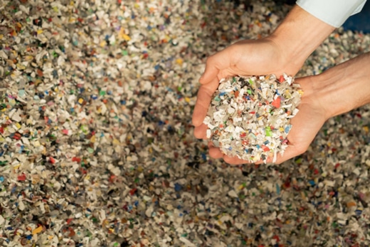 Dwie ręce trzymają porcję kolorowo wymieszanego recyklatu Ceramin®, a w tle widać dużą ilość materiału