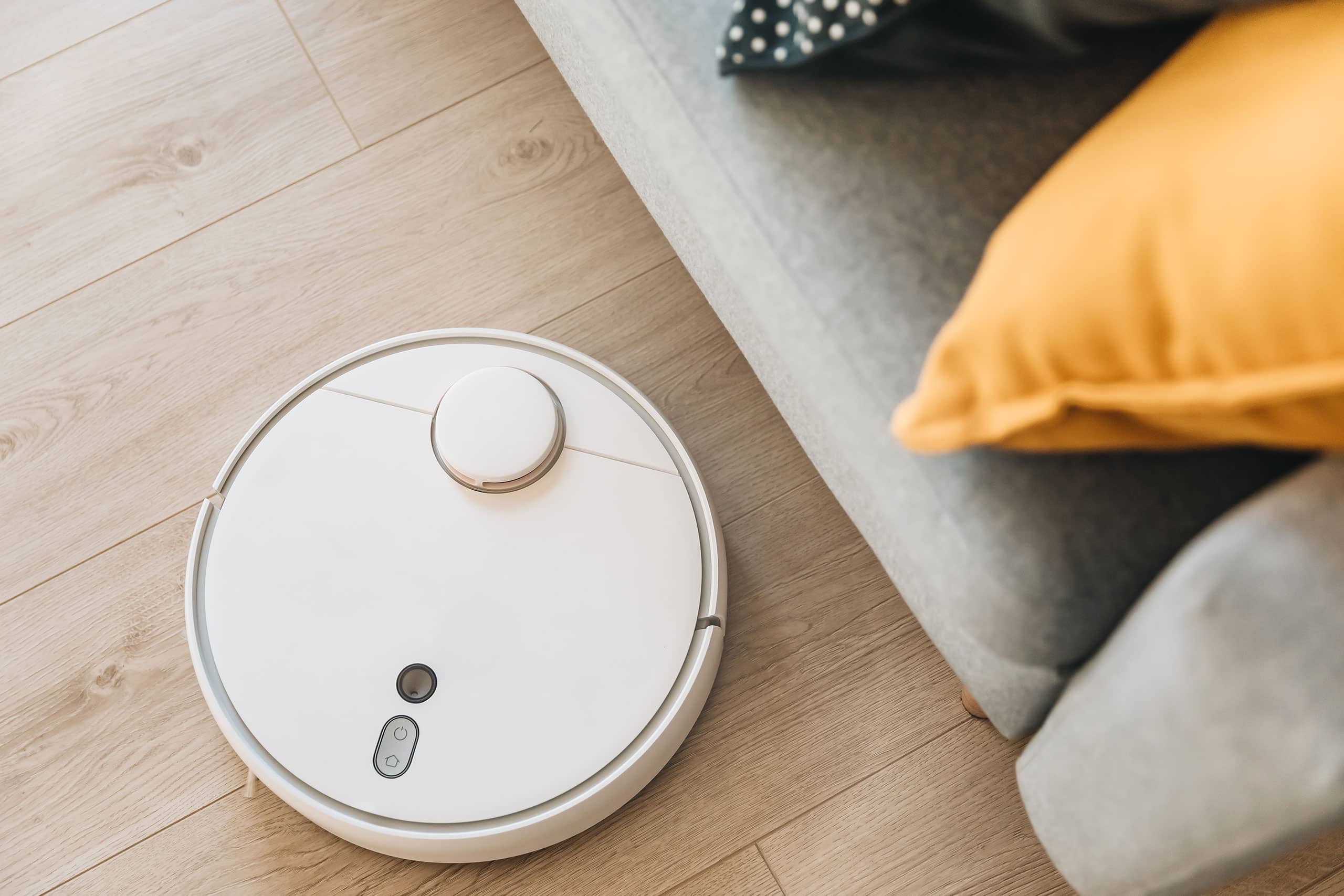 Biały robot odkurzający porusza się po jasnej, drewnianej podłodze obok szarej sofy z pomarańczowymi poduszkami.